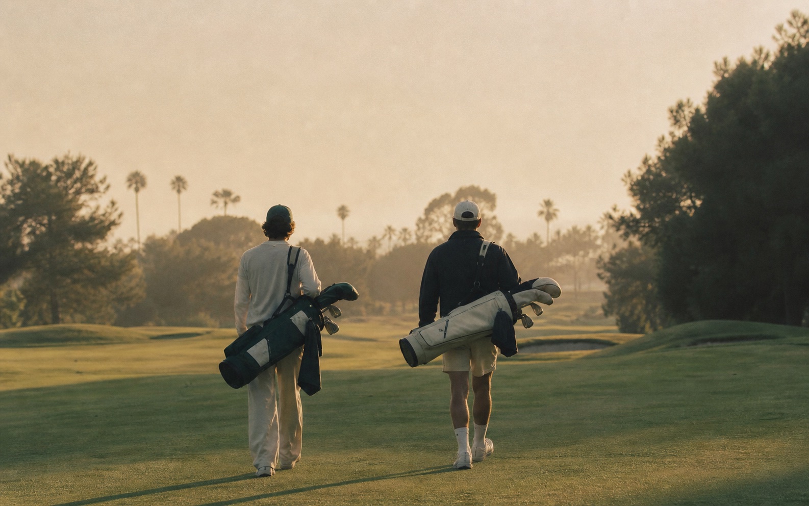 Two friends walking the fairway at golden hour, carrying golf bags.